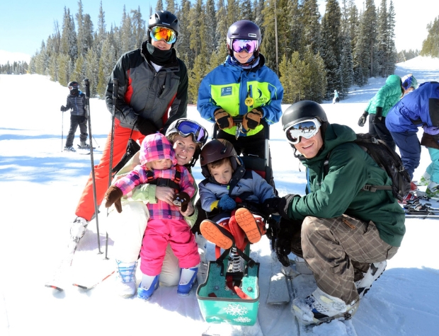 a young Ignite Athlete sitting in sit ski while his family and an Ignite Volunteer Instructor surrounds him smiling