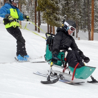 Ignite Athlete Heather on a sit ski navigating down a run with an Ignite Volunteer Instructor following behind