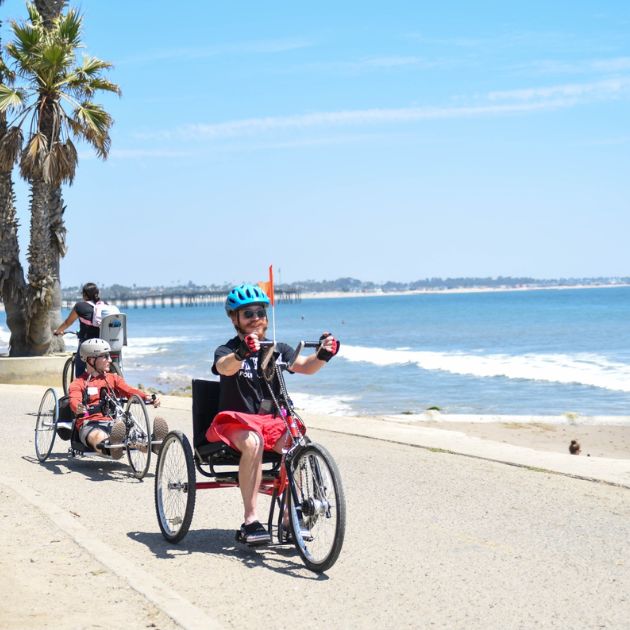 Two individuals using handcycles along a sunny beachside path.
