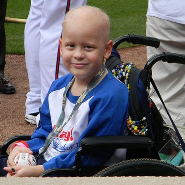 A young child in a wheelchair, smiling while holding a baseball.
