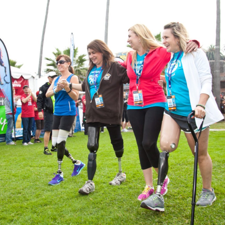 Four women, three with prosthetic legs, walk arm-in-arm with joyful expressions during a community event