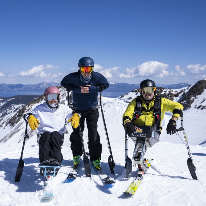 Three adaptive skiers, including two using sit-skis, smile atop a snowy mountain